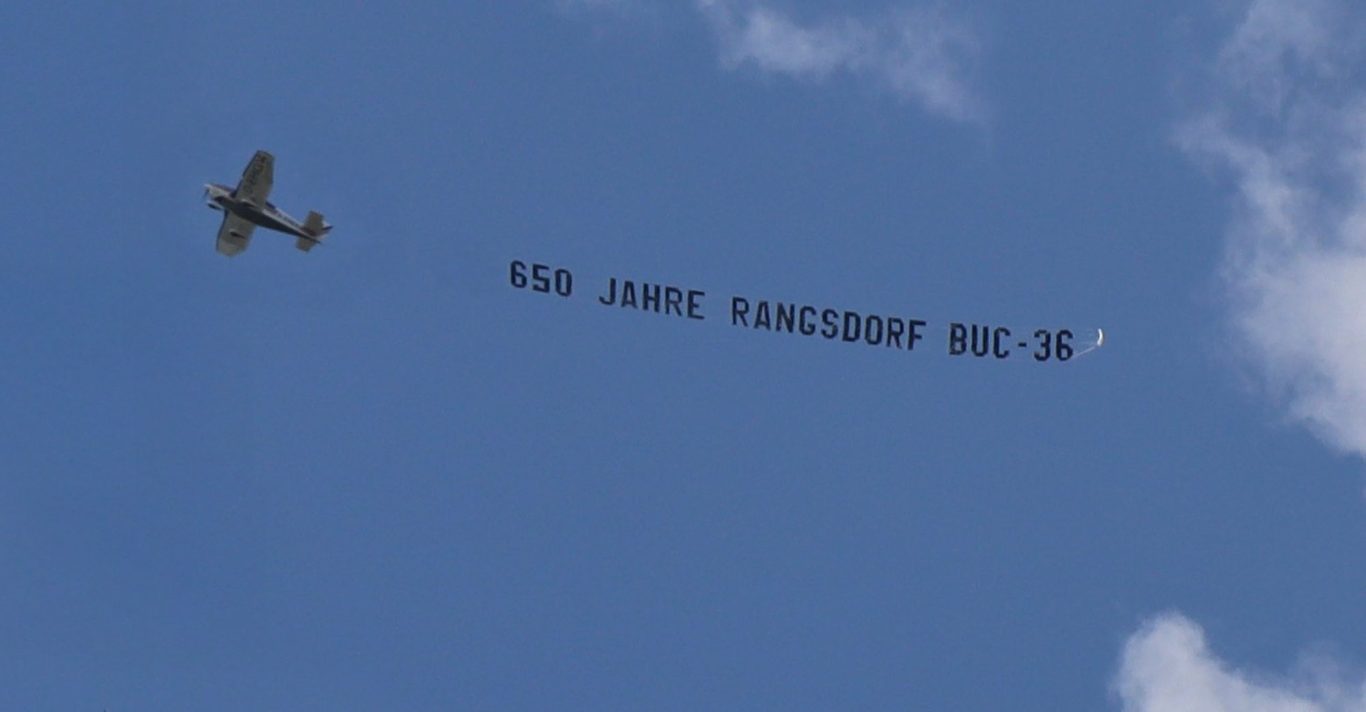 Ein Flugzeug zieht ein großes Banner mit der Aufschrift „650 Jahre Rangsdorf BUC-36“.